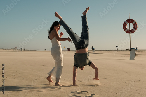 Man doing a handstand on a sandy beach while woman watches and laughs, playful and carefree romantic scene.
