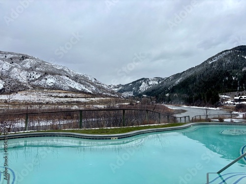 Outdoor hot spring pool overlooking a snowy river valley and mountains in Jackson Hole, Wyoming, with turquoise geothermal water contrasting winter landscape under overcast skies.