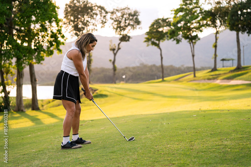 Hispanic woman focusing on her shot during a golf game on a lush green course with trees and water at sunset