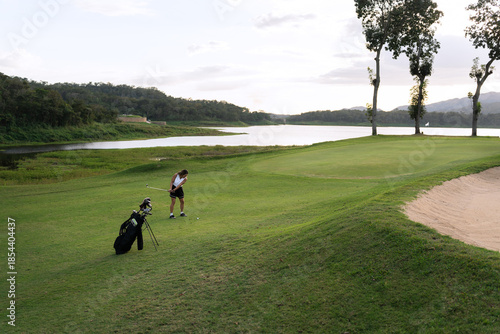 Woman swinging a golf club on a green course with a lake and trees in the background under a cloudy sky
