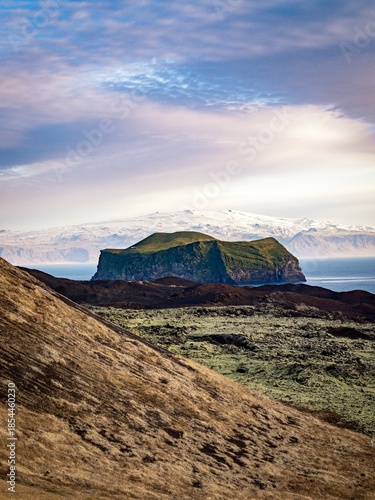 Iceland; Bjarnarey island, one of the Vestmannaeyjar, (Westman) islands, with  snow topped mountains of the mainland in the distance and dramatic sky. Isolated, uninhabited and known for its puffins.