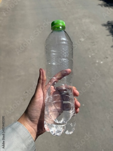 person's hand holding a clear, empty plastic water bottle with a bright green cap outdoors. The bottle features a hexagonal textured pattern.