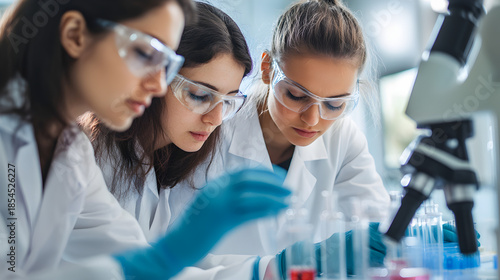 Three women in lab coats and safety goggles examining test tubes in a laboratory setting with microscope