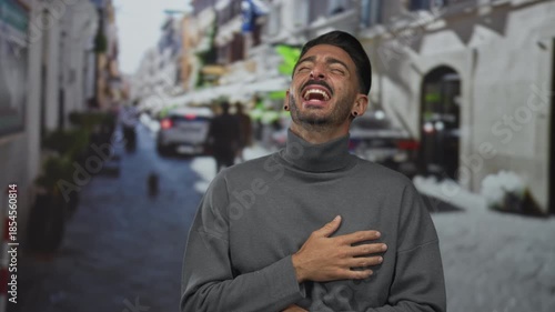 Man holds hand on chest and laughs broadly on narrow sunlit city street lined with blurred buildings; joy.