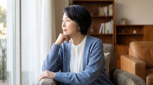 thoughtful middle aged woman sitting on living room sofa gazing out bright window in quiet home interior with soft natural daylight