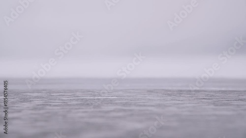 A close-up of drifting snow across the surface of a frozen lake. Wind blows snowflakes across the ice. Slow motion with shallow depth of field.