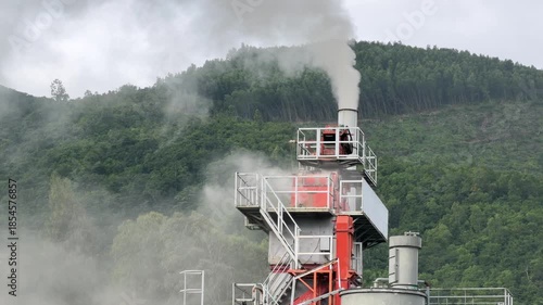 A factory smokestack releases thick smoke against green hills and cloudy sky, the slow drifting plume emphasizing environmental tension within a natural landscape. Industrial pollution.