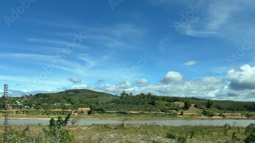 Blue sky above cultivated fields and rolling terrain shifts gradually with vehicle motion, producing a calm visual passage through open countryside and working land. Rural landscape.