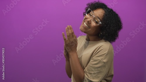 Woman wearing glasses and a beige shirt with clasped hands and curly hair smiling in studio; joy gratitude.