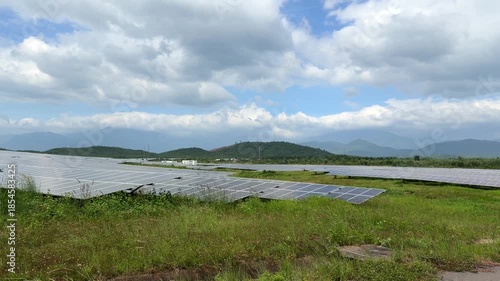 Rows of solar panels sweep across open ground under calm daylight, reflecting soft highlights bordered by distant hills and moving clouds. Sustainable living.