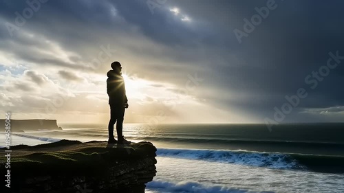 Lone figure standing on cliff edge overlooking stormy ocean waves.