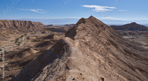 A sharp mountain ridge rises above wide and open valley of Nahal Zinin or Zanin in the Negev. A pair of tourists stands on a cliff with a panoramic view of the valley. Panoramic desert landscape.