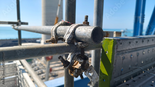 The metal scaffolding pipes and couplers secured with bolts at an industrial construction site, with offshore platform and blue sky in background, highlighting structure, safety and engineering detail