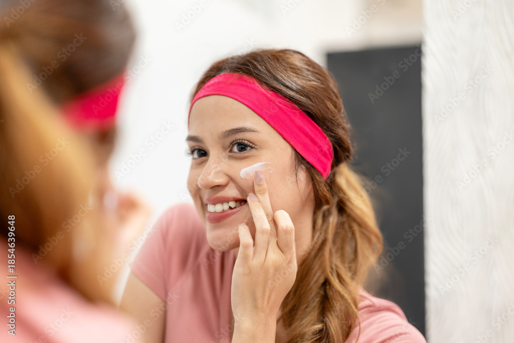 Fototapeta premium Indonesian southeast asian woman wearing a pink headband applies facial cream to her face while looking in the mirror at home. Doing her daily skincare routine and enjoying a moment of self care