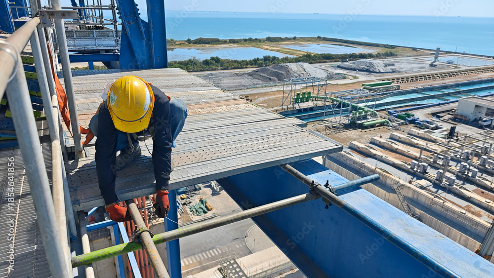 Fototapeta premium Construction worker wearing helmet and safety gear installing scaffolding at height on an industrial site near the sea, emphasizing teamwork, safety procedures, and offshore engineering environment.