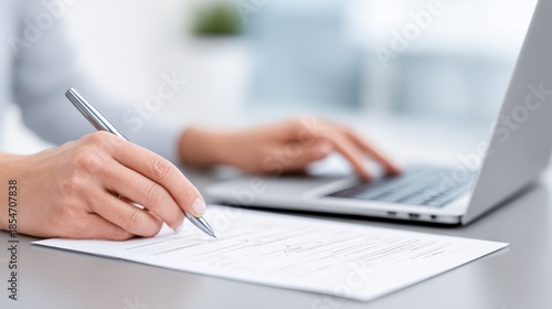 Professional accountant, a woman with light skin, is writing on a document while using a laptop, showcasing a modern workspace with focus on financial tasks