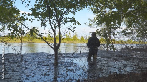 A fisherman stands in the bushes in the water and fishes with a fishing rod. Dense tree branches frame the scene, creating a natural, serene riverside environment. Camera panning