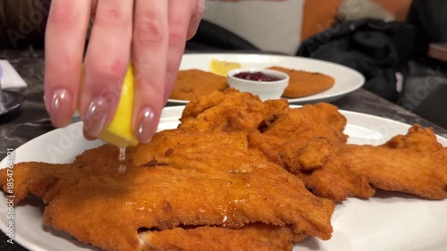 pour lemon juice over the schnitzel Crispy chicken schnitzel served on a gray ceramic plate with a lemon slice and fresh herbs on a white wooden table.