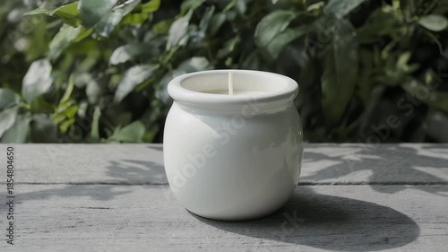 Wallpaper Mural A white ceramic candle holder, centered on a light gray wooden surface. Lush green foliage forms a soft backdrop. The candle inside is white Torontodigital.ca