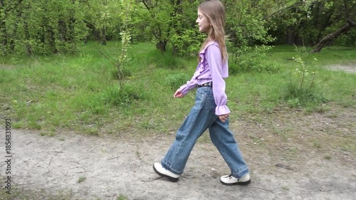 A young girl with long hair walks along a dirt path in a green park, wearing a purple top and jeans, enjoying a peaceful day outdoors.