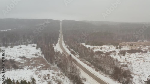 Drone view showing a single car traveling on a long, straight road through a vast, snow-covered forest on a cold and foggy winter day