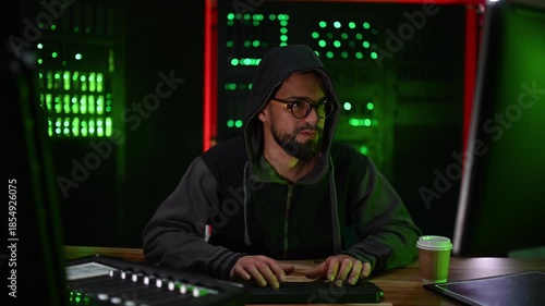 Hooded hacker smiling while typing on keyboard in a dark server room