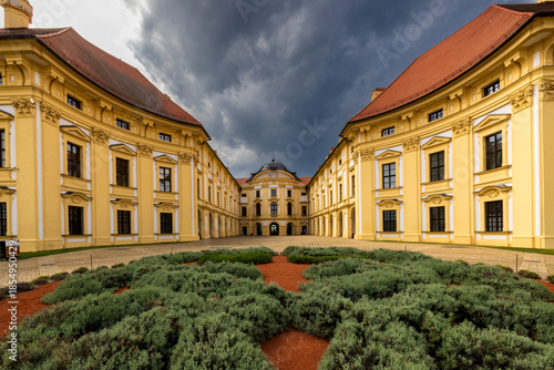 Grand yellow baroque palace courtyard under a dramatic cloudy sky.