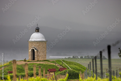 Stone chapel on a green hill overlooking a vineyard under a foggy sky.