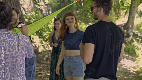 A joyful group of young friends collaborates to set up a bright green hammock among the trees in a sunny forest. They are enjoying a relaxing day outdoors, sharing smiles and good company.
