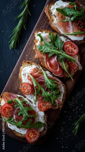 Gourmet open faced sandwiches with prosciutto, fresh arugula, creamy soft cheese and cherry tomatoes served on toasted artisan bread on a rustic wooden board in dark moody food styling