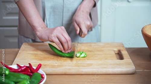Hands slicing green and red chili pepper on wooden cutting board in home kitchen