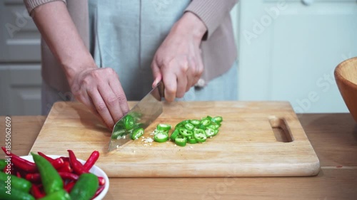 Hands slicing green and red chili pepper on wooden cutting board in home kitchen