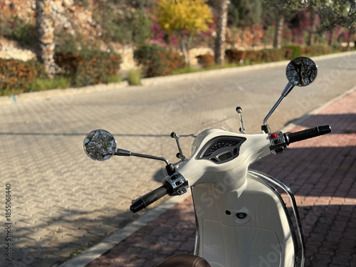 Cream-colored scooter parked on sunlit cobblestone street with blurred trees in background