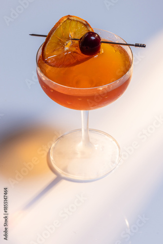 Beautiful amber cocktail, a Negroni or manhattan served up on a white surface with dramatic lighting.