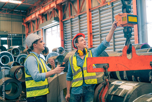 Caucasian industrial engineer men controlling overhead crane with remote in factory. Worker partner wear safety helmet vest lifting heavy metal coil material. Manufacturing hoist machine logistics