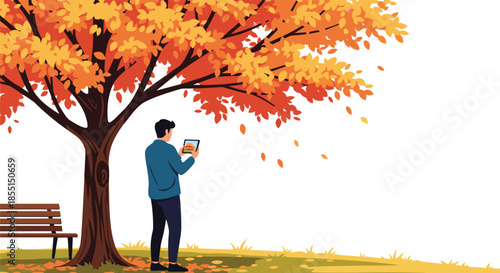 Man standing under a large tree with vibrant orange leaves taking a photo with his tablet in a peaceful autumn park.