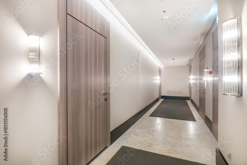 A hallway is visible with light fixtures on the walls. Two doors are seen on one side. The floor is covered with a carpet and the walls are painted in light colors