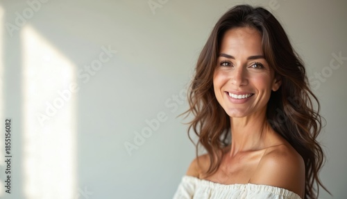 Free Download Middle age woman with long wavy brunette hair smiles gently. Natural makeup, soft expression, elegant off shoulder top. Skincare beauty confidence, serene peaceful background. - GFXLoad