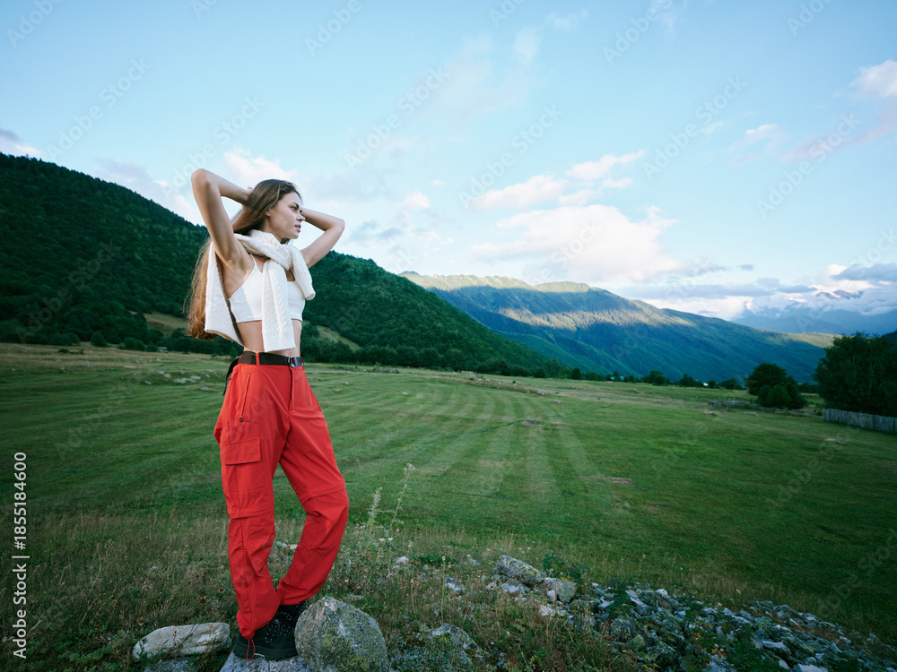 Naklejka premium Outdoor landscape field mountains, a woman standing in a grassy plain wearing a white top and red pants, with hands behind head, enjoying expansive space and fresh air under a bright sky.