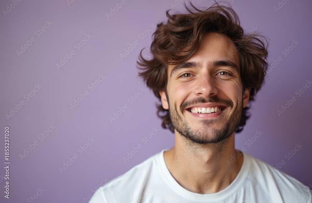 Fototapeta premium Young man with brown wavy hair smiles happily. He wears a white t-shirt. Plain purple background emphasizes his cheerful face. His eyes sparkle with joy and confidence.