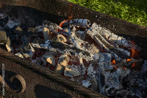 Smoldering logs and bright flames in a metal barbecue grill. Detailed close-up of fire, heat, and smoke outdoors. Flaming logs with heat and smoke rising from an open grill.