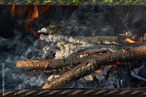 Smoldering logs and bright flames in a metal barbecue grill. Detailed close-up of fire, heat, and smoke outdoors. Flaming logs with heat and smoke rising from an open grill.
