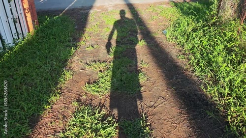 The setting sun casts a long shadow of a man walking along a grassy path that eventually meets a sidewalk. The scene is captured from a first person perspective