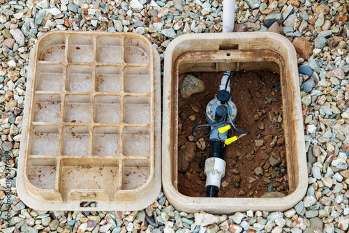 Opened ground-level sprinkler control box holding connecting pipes and electric valve with its lid placed next to it