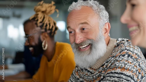 A man with a beard and glasses is smiling at the camera. He is surrounded by other people, including a woman with a pinkish face. Scene is friendly and welcoming