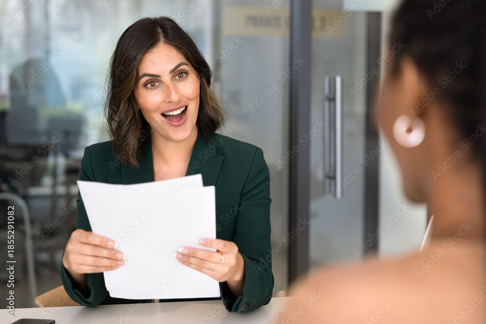 Obraz premium Excited positive HR manager woman meeting with job applicant at office table, holding paper documents, resume, learning about spectacular professional skills of candidate, gasping in surprise