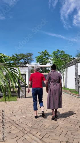 Lilongwe Malawi Africa, 01.12.2025. Video. African female  carer going for a walk in a courtyard with an elderly Dementia sufferer  on holiday in Malawi Africa.