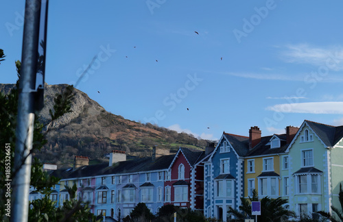 Colorful Victorian houses line a street under a bright blue sky with distant mountains, bathed in sunlight, creating a cheerful, picturesque scene in Llanfairfechan - Wales - UK

