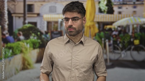 Young hispanic man wearing beige shirt covers face with hands on sunlit city street by cafe seating and bicycles; embarrassment.