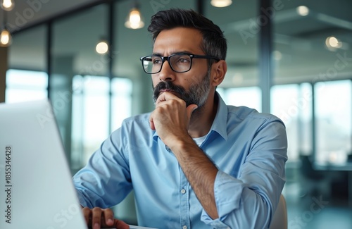 Man in glasses ponders project at desk with laptop. Indian pro thinks deeply, considers options for startup growth in modern office setting. Focused businessperson plans strategy. Digital world.